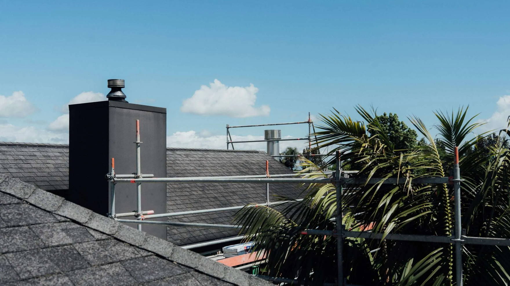 Scaffolding erected around a Tauranga house with a dark chimney and palm trees behind