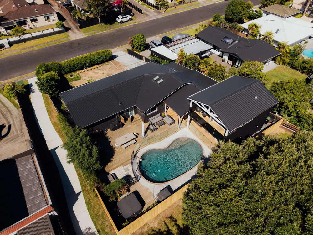 Aerial view of a dark roofed Tauranga home with a curved pool and a separate cabin in the garden