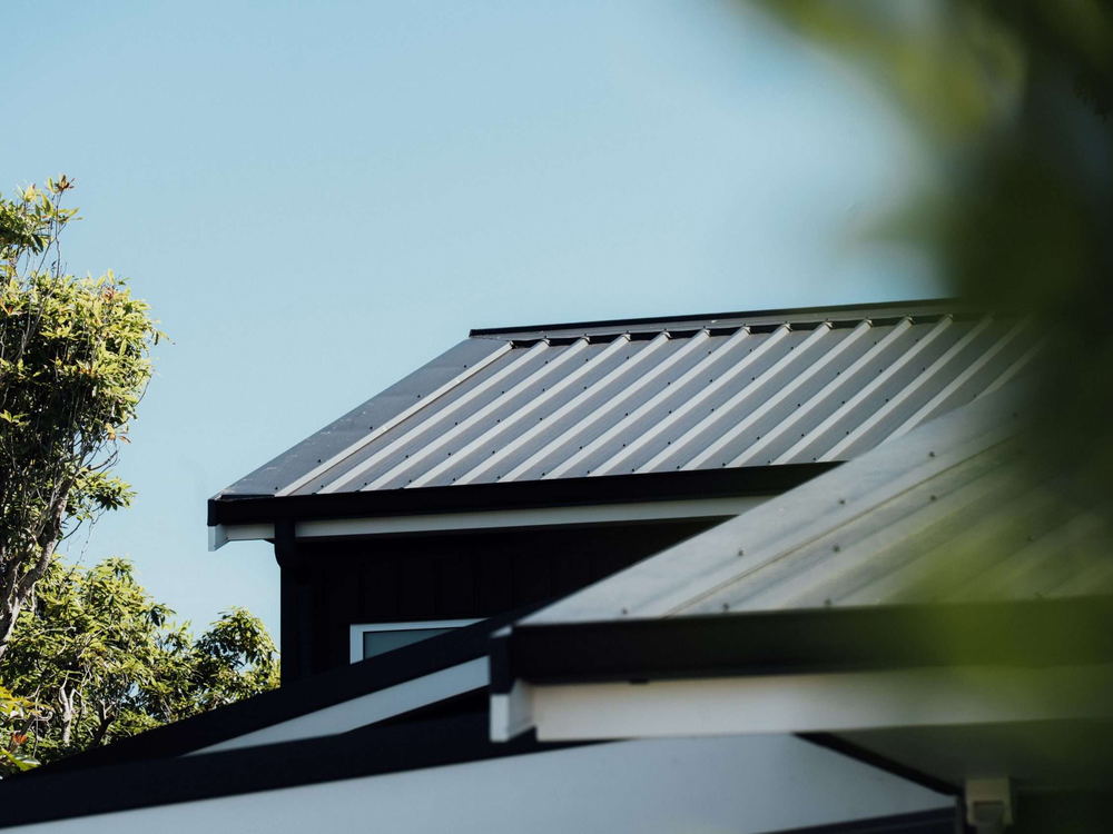 Close view of a dark metal roof edge and barge flashing against a bright sky and trees
