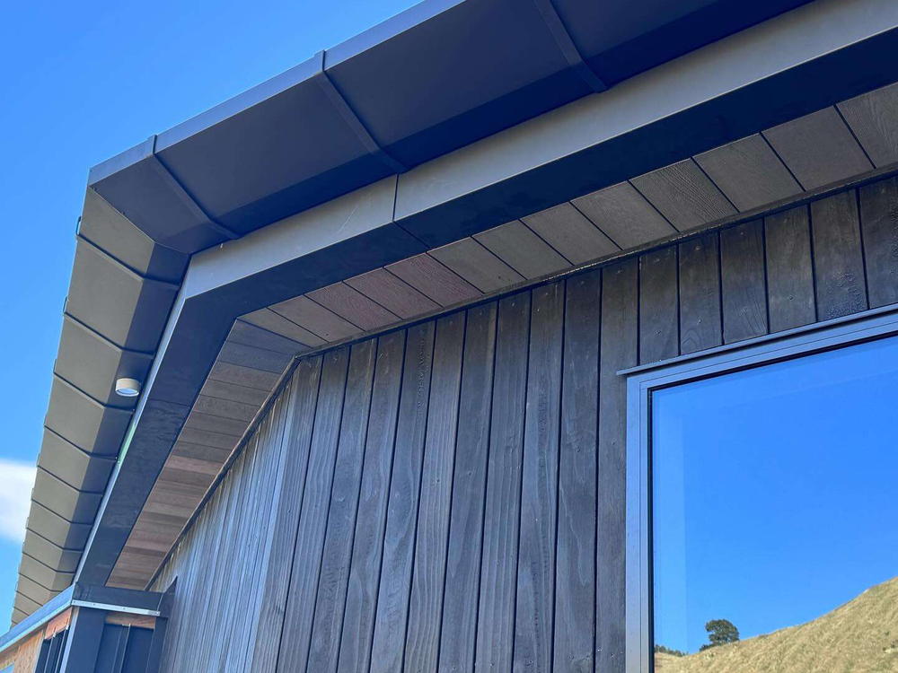 Dark tray roof and barge meeting vertical timber cladding, with farmland reflected in the glazing