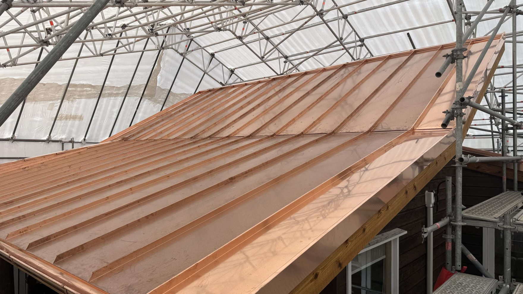 Copper architectural tray roof being laid on a curved roof under a shrink-wrapped scaffold