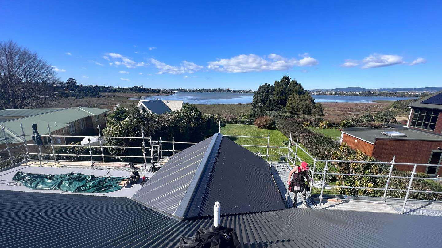 Dark longrun steel roof running down to the ridge with roofers working on it and the estuary behind